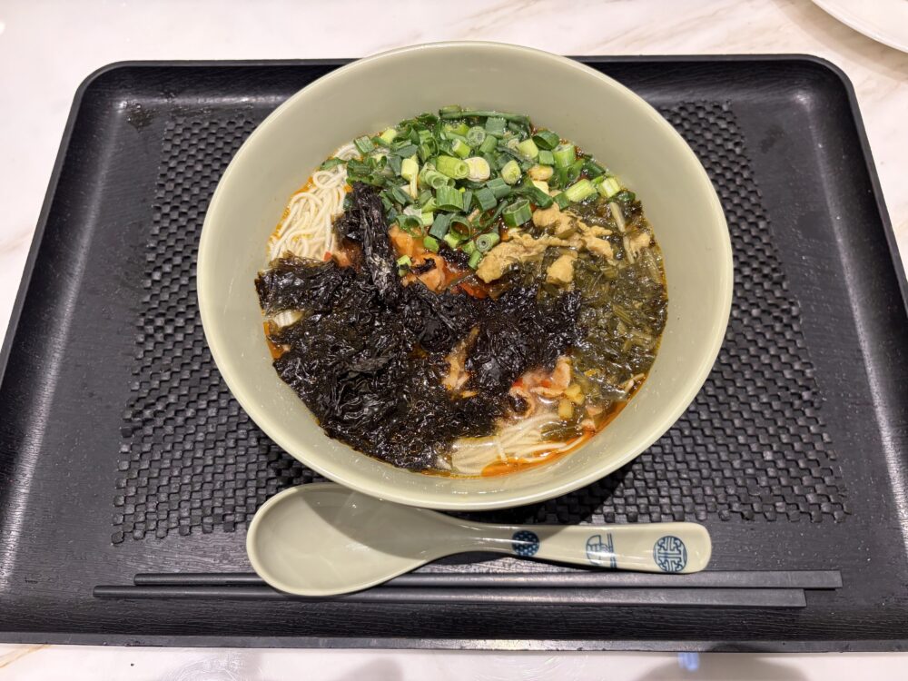 上海浦東国際空港V39ラウンジのヌードルバーで提供される出来立てのラーメン。黒いトレーに乗った器に、海苔、刻みネギ、高菜などのトッピングが盛り付けられ、ラー油の浮いたスープと細麺が写っている様子